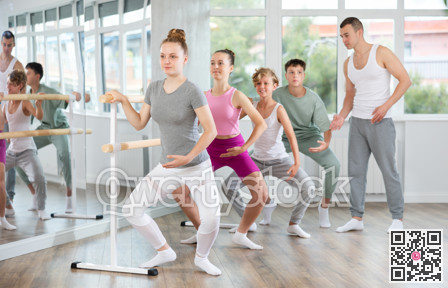 Male teacher conducts class with teens in ballet studio, observes doing plie exercise.