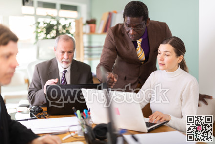 African man leaned over to seated female employee, helps with work and looks at laptop screen