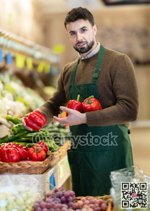 Young male seller putting red bell peppers on counter in grocery market