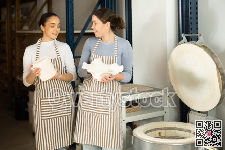 Two girls craftperson stand at furnace and brag to each other about finish clay products