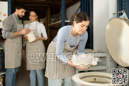 Two female potters take finished ceramic dishes, various mugs and a vase out of oven