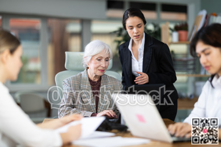 Young female leaned over to seated senior woman employee, helps with work and looks at laptop screen