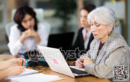 Tired mature woman is sitting at computer at work in crowded office