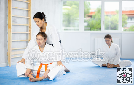 Young woman in kimono stretching in butterfly pose with assistance of trainer