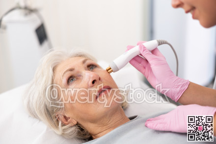 Young beautician doctor doing RF lifting procedure for face lifting of mature woman laying in beauty salon. Hardware cosmetology