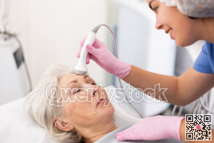 Professional young female cosmetologist doing facial skin ultrasound cavitation for for face lifting of mature woman laying in beauty salon