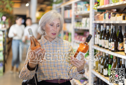 Senior female customer in supermarket examines bottles of wine, chooses alcoholic drink.