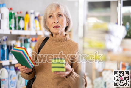 Elderly housewife chooses sponges for washing dishes and tiles in supermarket
