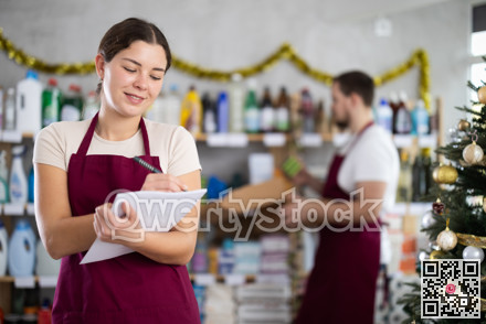 Attentive saleswoman restock of goods, checking papers, male assistant laying out goods in background in small household store decorated for New Year celebratio