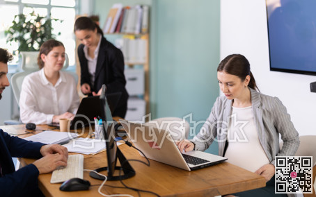 Pregnant woman is working on a laptop in an office while colleagues are discussing a project