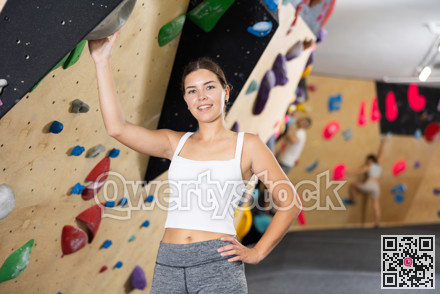Positive slender girl in sportswear is standing near climbing wall in gym