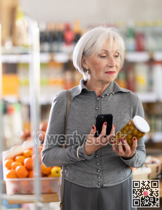 Checking expiration date of canned green olives - elderly female shopper scans QR code on label of jar using smartphone