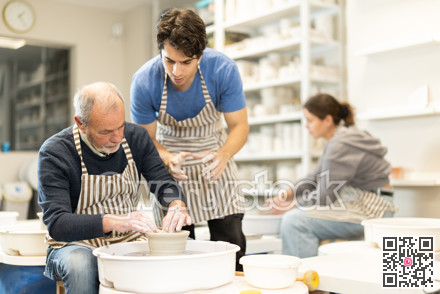 Young guy helping elderly man shape pottery on wheel
