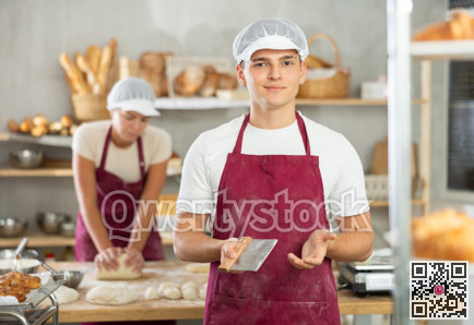 Young baker guy is standing near work table in kitchen, working with dough
