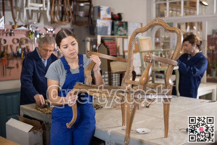 Skilled female furniture repair specialist in uniform restoring Victorian chair during workday in workshop
