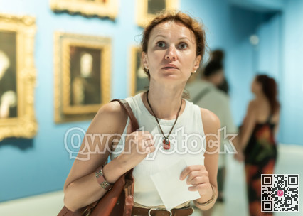 Interested woman looking closely at paintings in art gallery and holding tour sheet in hands