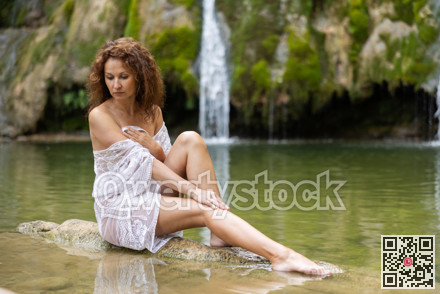 Sexy woman in white transparent mesh cape sits on rock on shore of waterfall